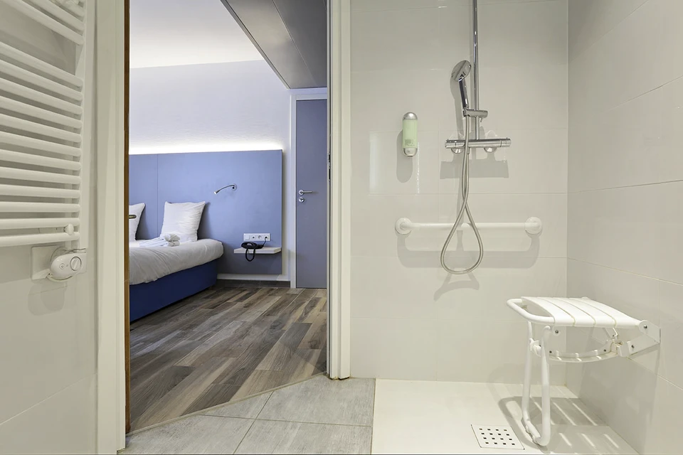 Salle de bain adaptée PMR avec douche à l'italienne équipée de barres d'appui murales blanches, siège de douche rabattable et tabouret blanc réglable